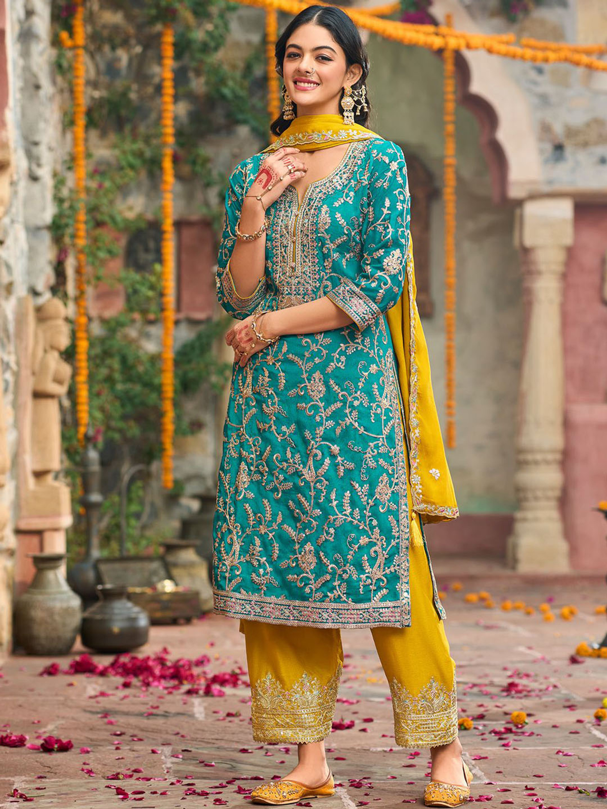 A woman models a traditional Indian salwar kameez suit in a courtyard setting. The outfit features a teal green embroidered tunic (kameez) with gold floral patterns, matching yellow pants (salwar), and a yellow dupatta.