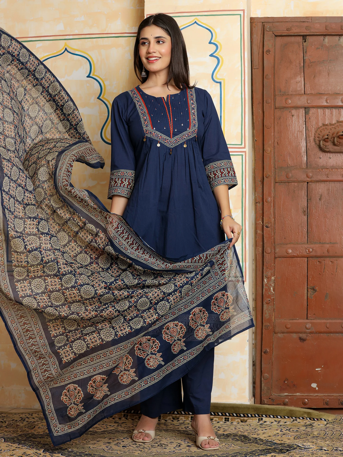 Woman holding a navy blue and beige patterned dupatta in front of a decorative wall and wooden door.