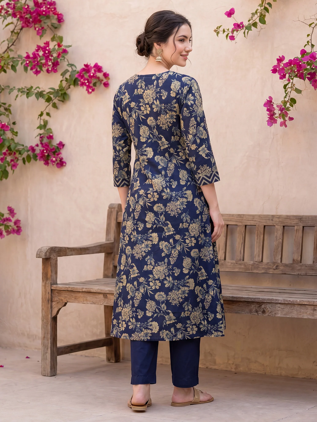 Woman wearing a blue floral kurta with matching pants standing in front of a wooden bench and pink flowers.