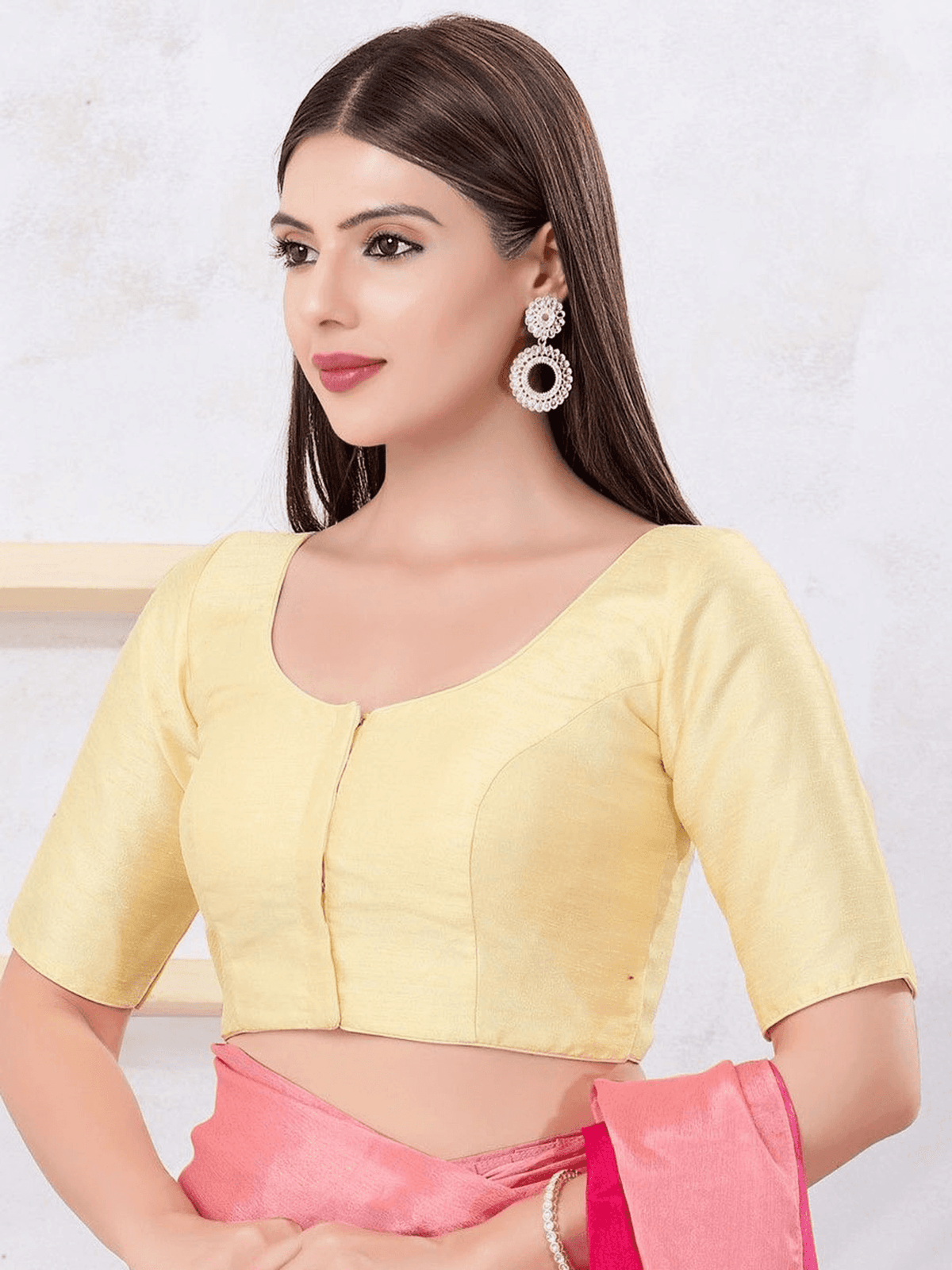 A woman modeling a pale yellow short-sleeve blouse with a scoop neckline and front button detail, paired with a pink skirt. She is wearing large silver-toned earrings against a light textured wall background.