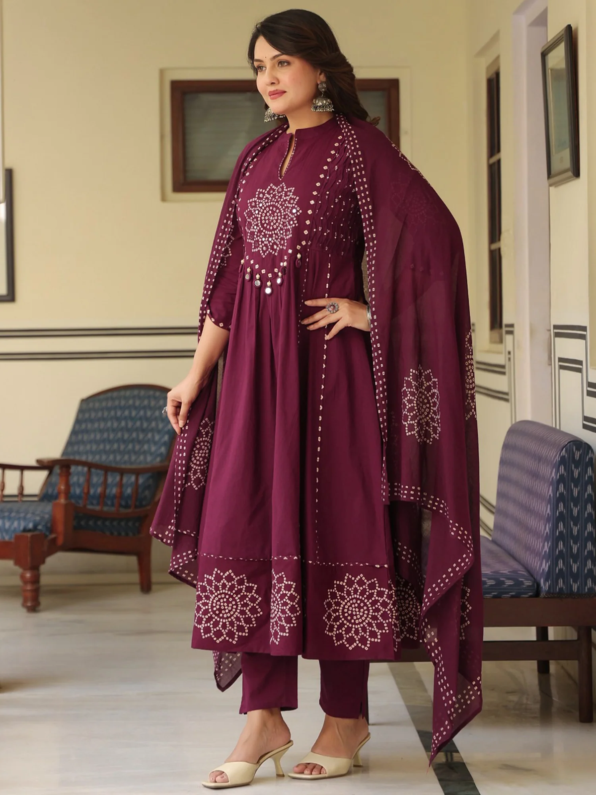 Woman in a maroon traditional outfit with intricate patterns in a room with chairs and a mirror.
