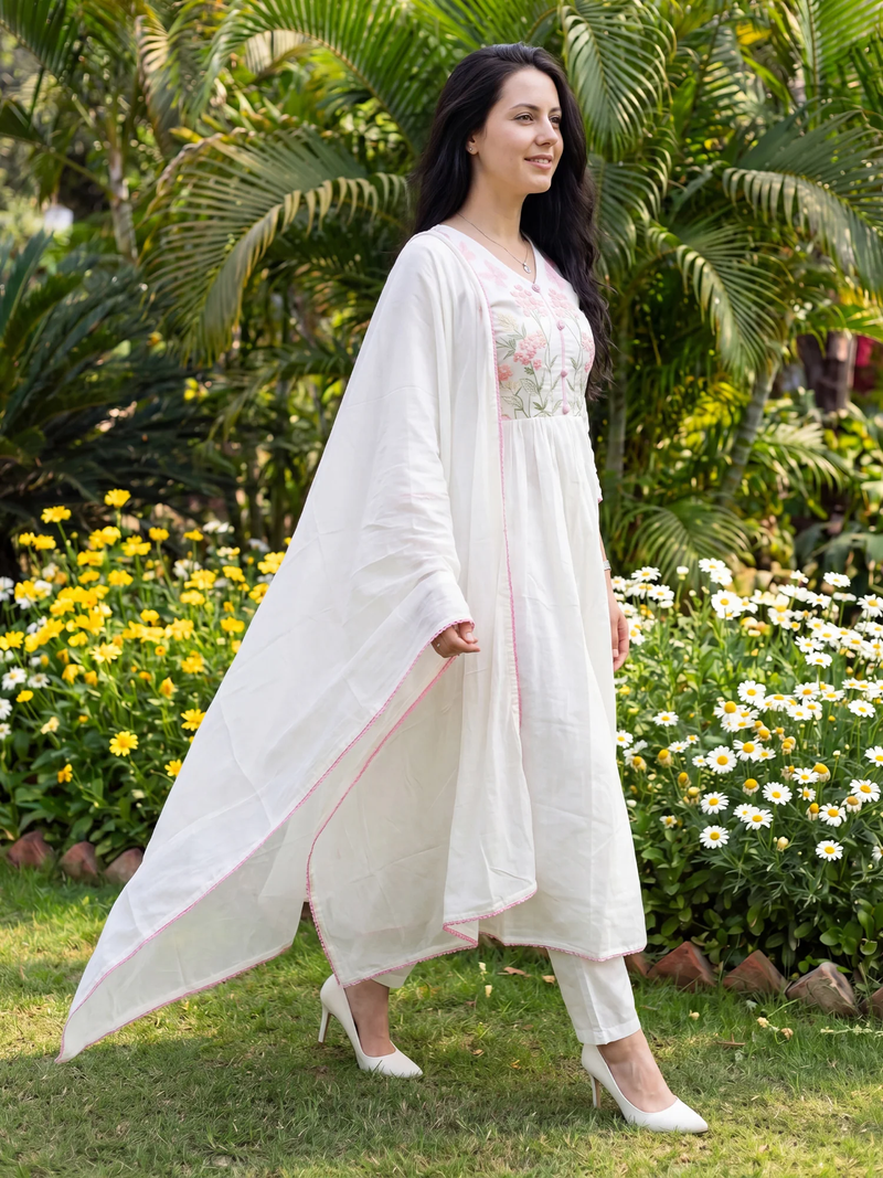 Woman in a white outfit standing in a garden with flowers and greenery.