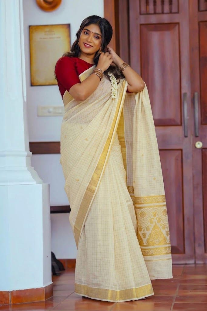 A woman wearing a traditional Indian saree with a cream-colored body, gold border, and a red blouse, standing in an indoor setting with wooden doors and a white pillar.