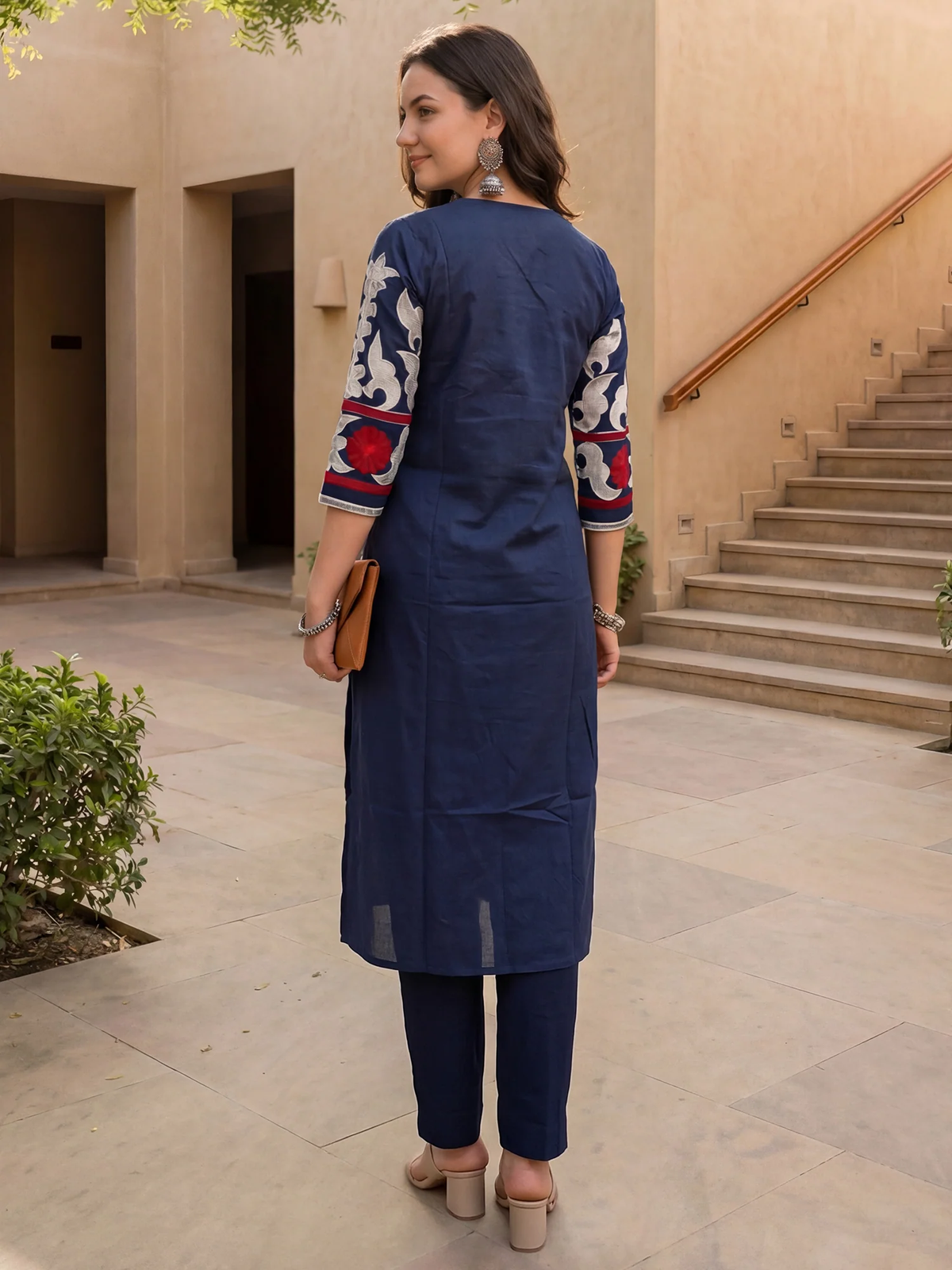 A woman wearing a navy blue kurta with red and white patterned sleeves, paired with matching navy trousers, standing in an outdoor courtyard.