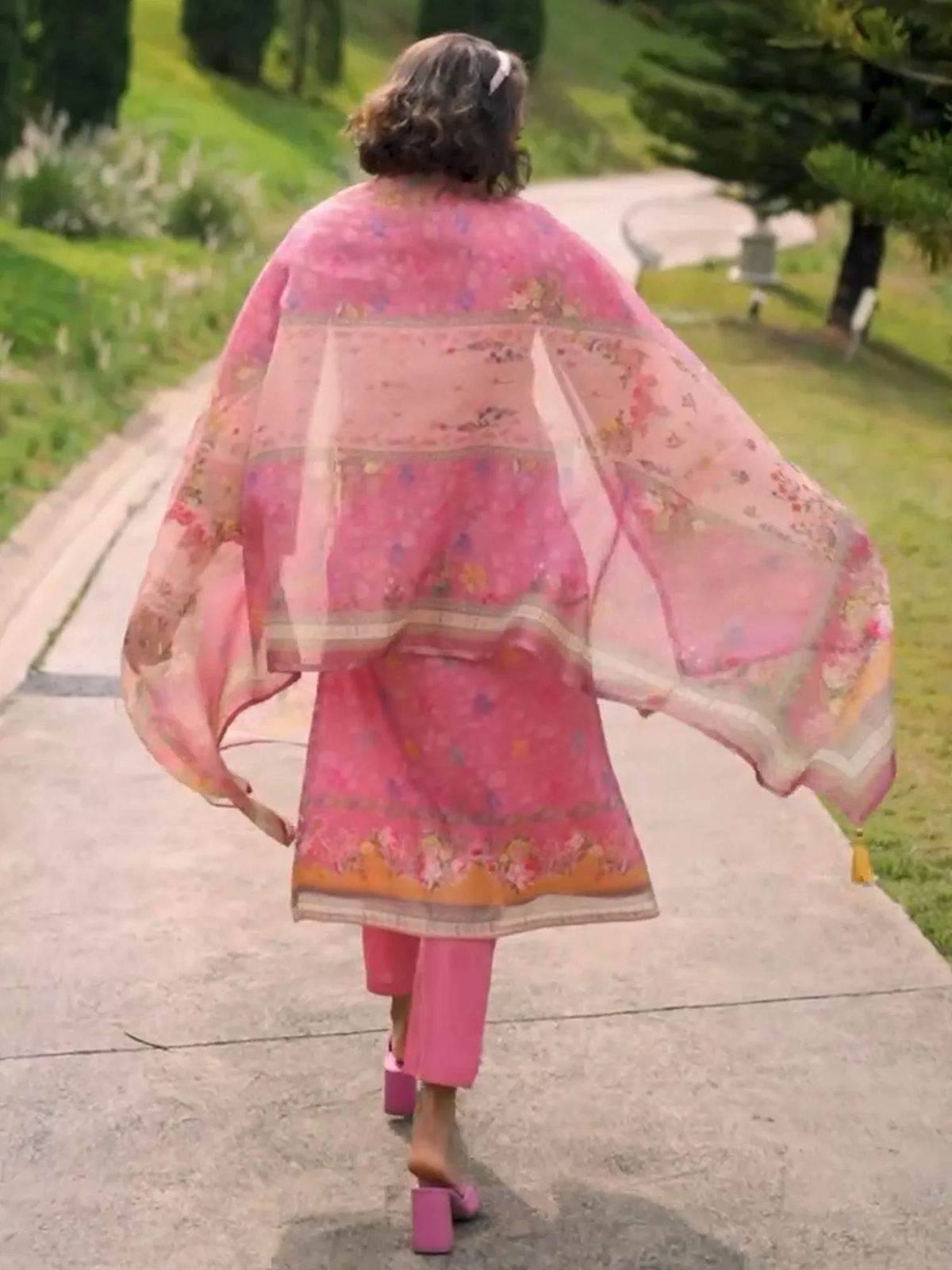 A woman viewed from behind walking on a paved path outdoors, wearing a pink traditional outfit with a sheer floral dupatta and pink high heels.