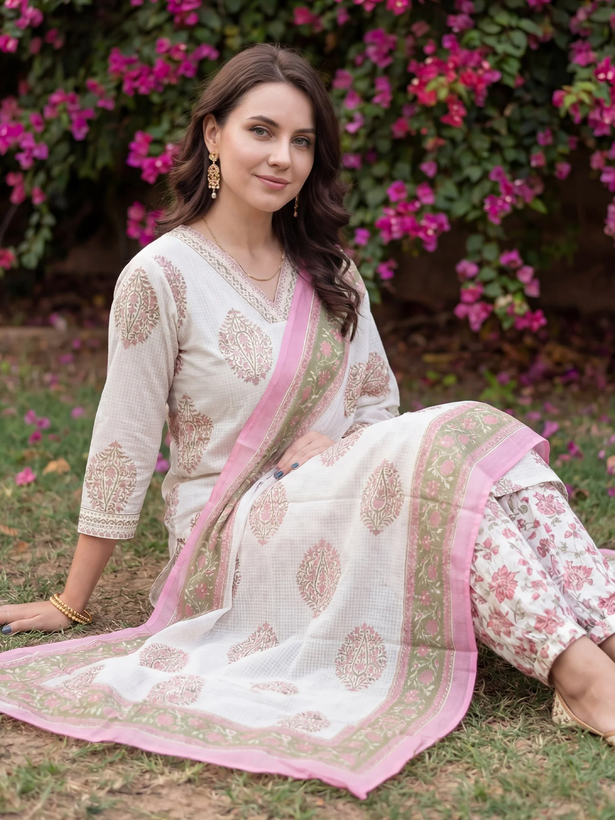 Woman in a white and pink traditional outfit sitting on grass with pink flowers in the background