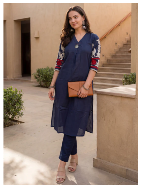 A woman modeling a navy blue kurta set with three-quarter sleeves featuring red and white floral embroidery, paired with matching navy pants, holding a brown clutch bag in an outdoor courtyard setting.