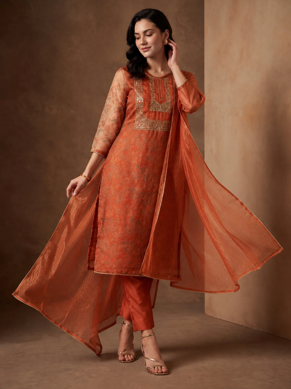 Woman wearing an orange traditional outfit with a sheer dupatta against a brown background