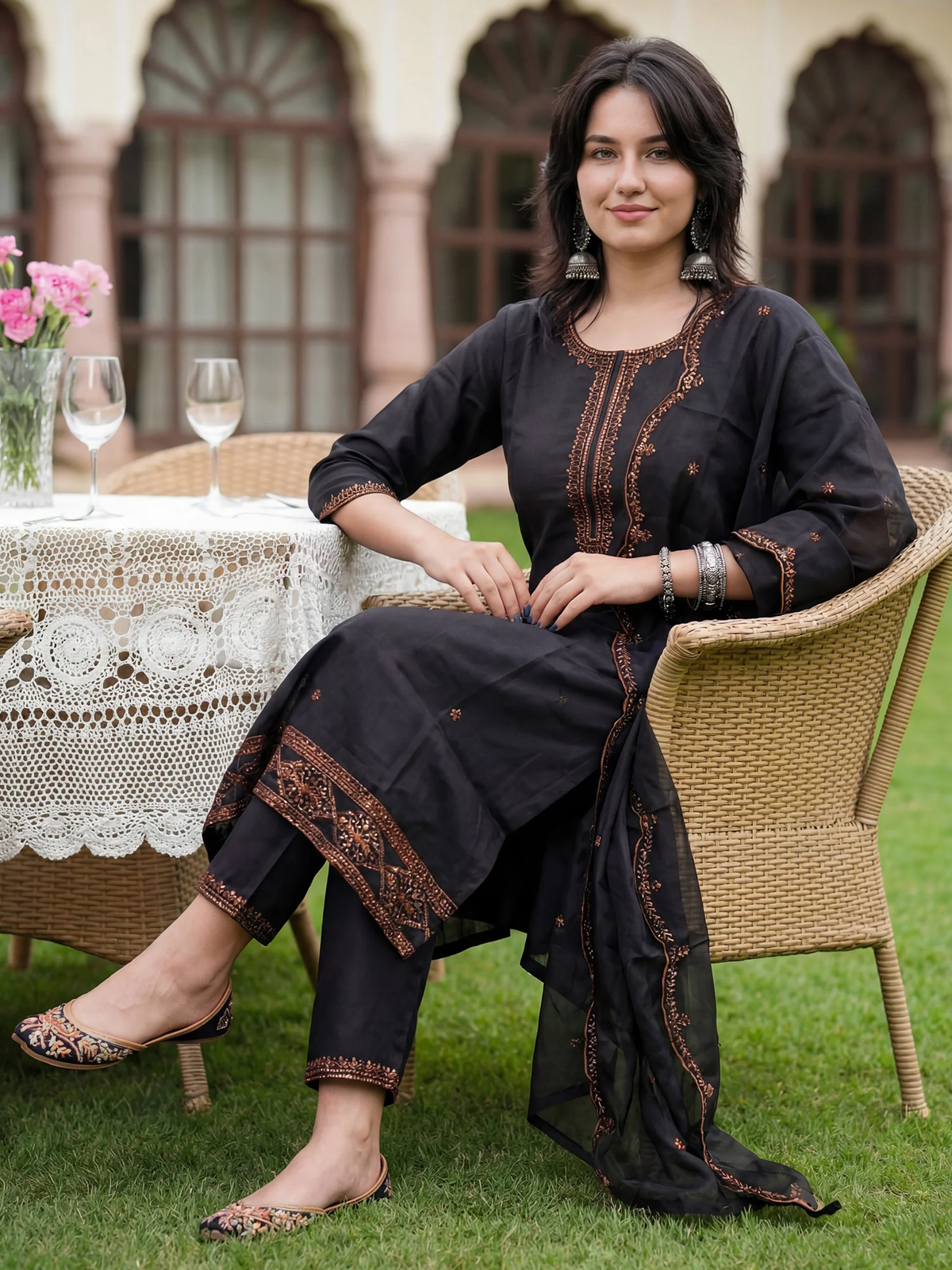 Woman in black embroidered outfit sitting outdoors on a grassy area with a table and chairs in the background.
