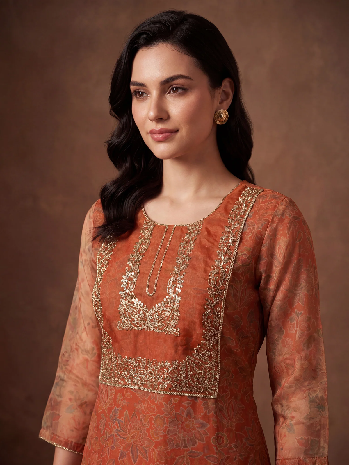 Woman wearing a traditional orange and gold embroidered outfit against a brown background