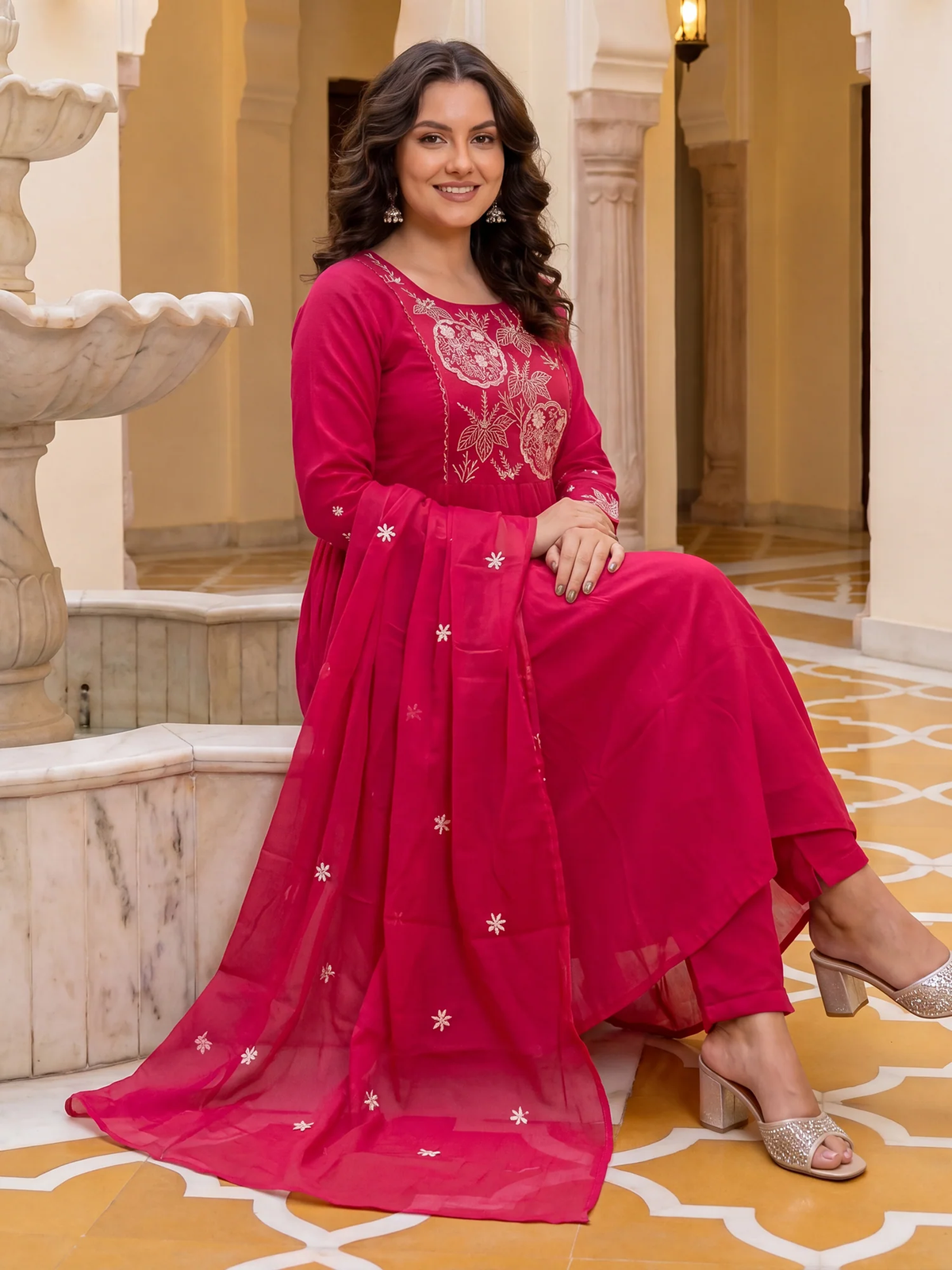 Woman in a pink traditional outfit with intricate designs sitting in an elegant interior setting.