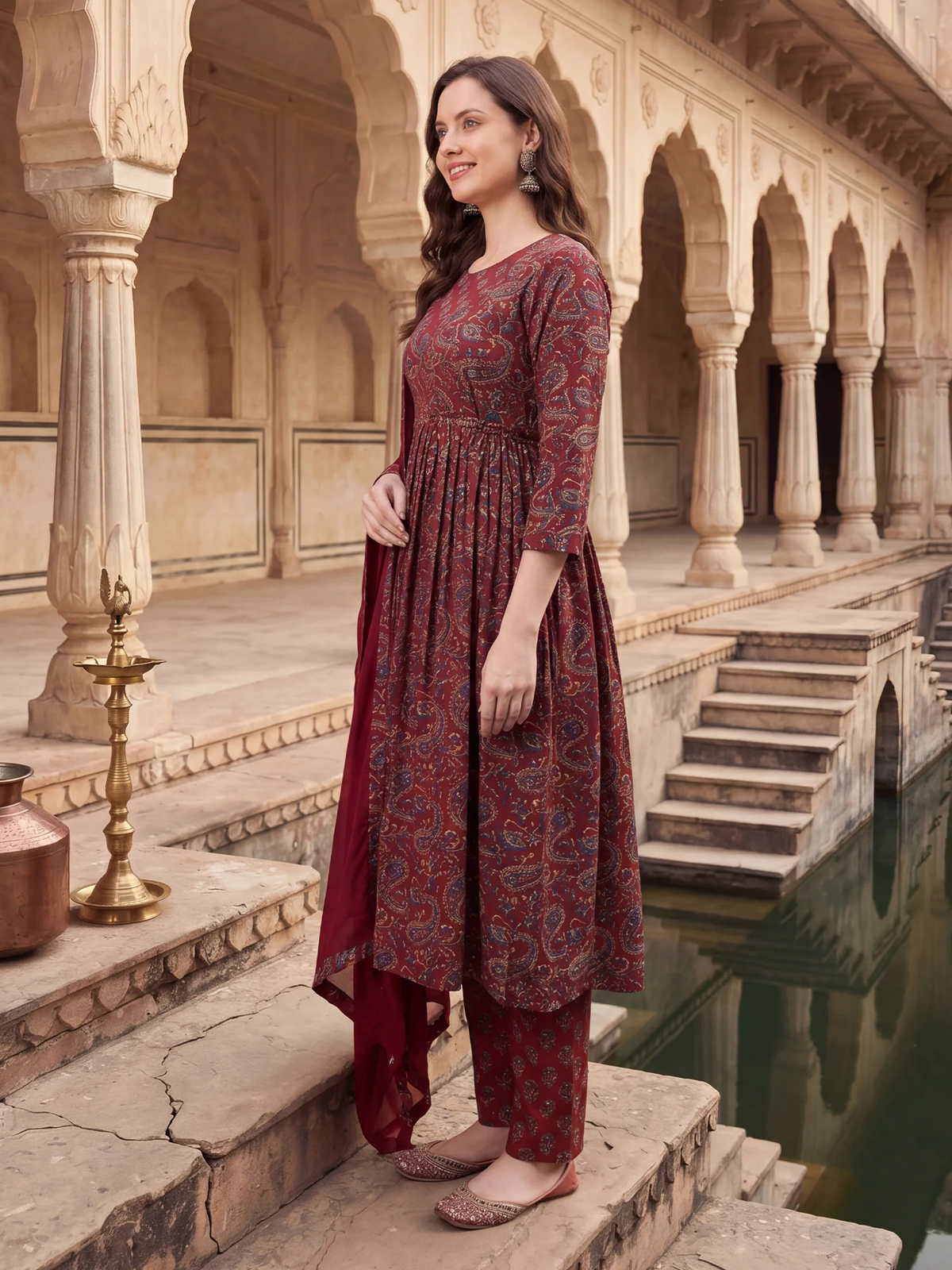 A woman modeling a traditional Indian salwar kameez set in a courtyard with arches and a reflecting pool. The outfit features a deep red base with intricate paisley patterns in blue, gold, and brown.