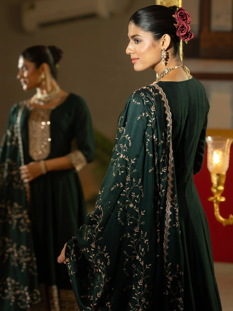 Woman in a dark green traditional outfit with floral patterns, standing in a softly lit room.