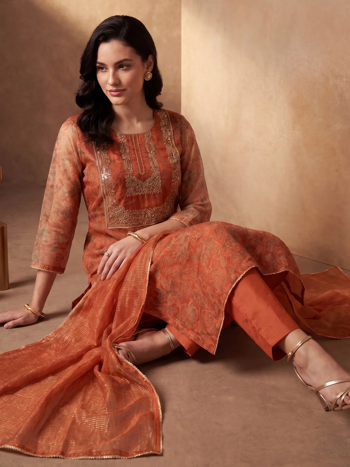 Woman wearing an orange traditional outfit with intricate patterns sitting on a beige floor.