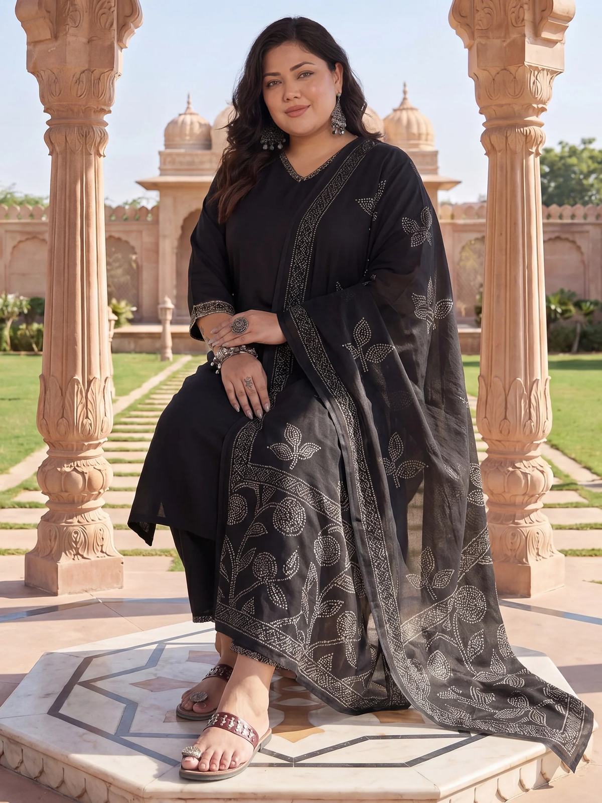 Woman in a black traditional outfit with a floral patterned scarf standing in an outdoor setting with architectural columns.