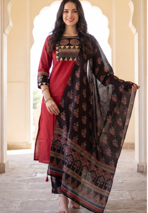 A woman modeling a traditional three-piece salwar kameez set in a corridor with arches. The set features a red kurta with geometric embroidery at the neckline, matching red trousers, and a black dupatta with red and gold floral prints and borders.
