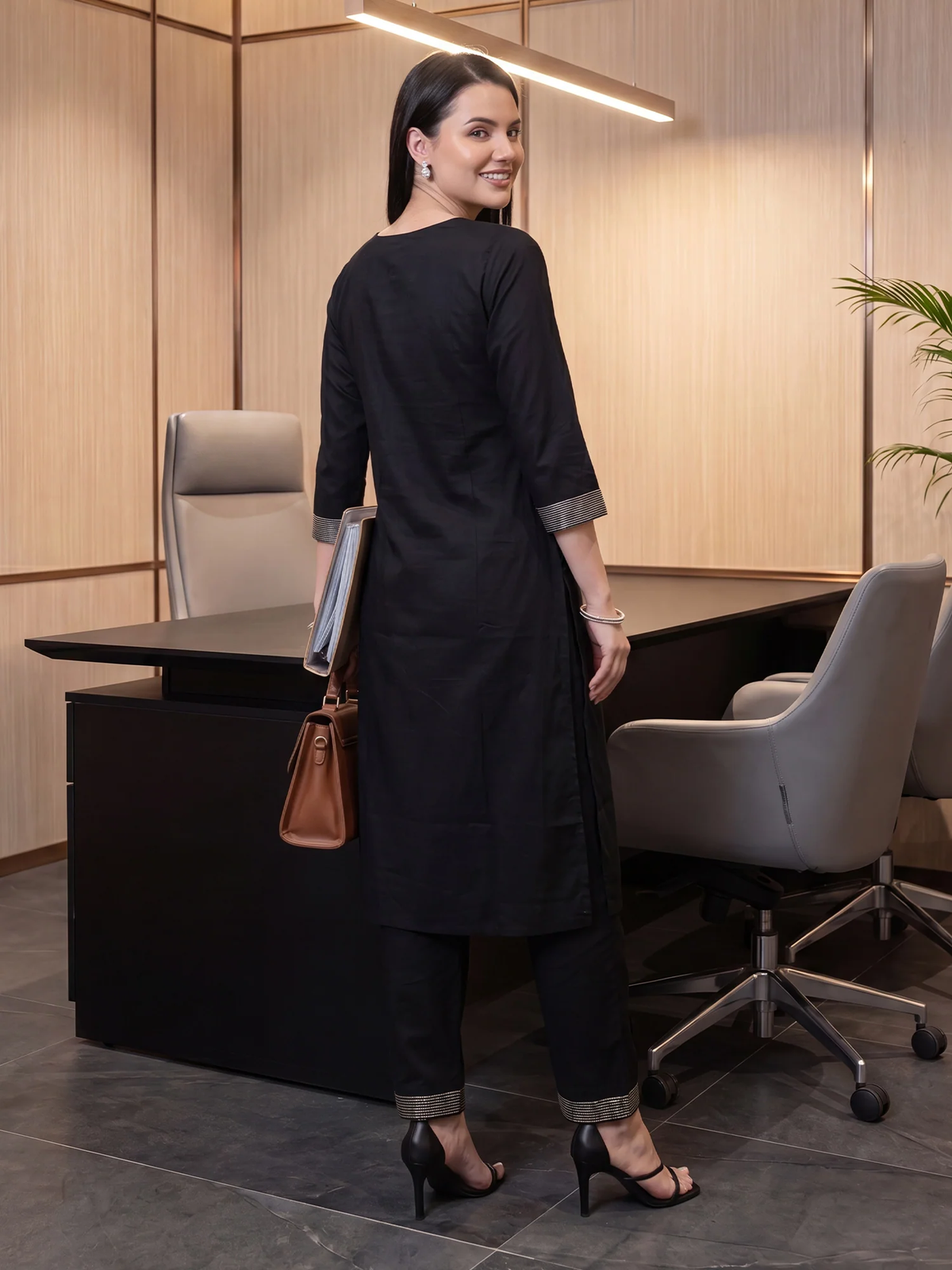 A woman wearing a black three-quarter sleeve kurta with gray rolled cuffs and matching black trousers, standing in a modern office setting with a desk and chairs.