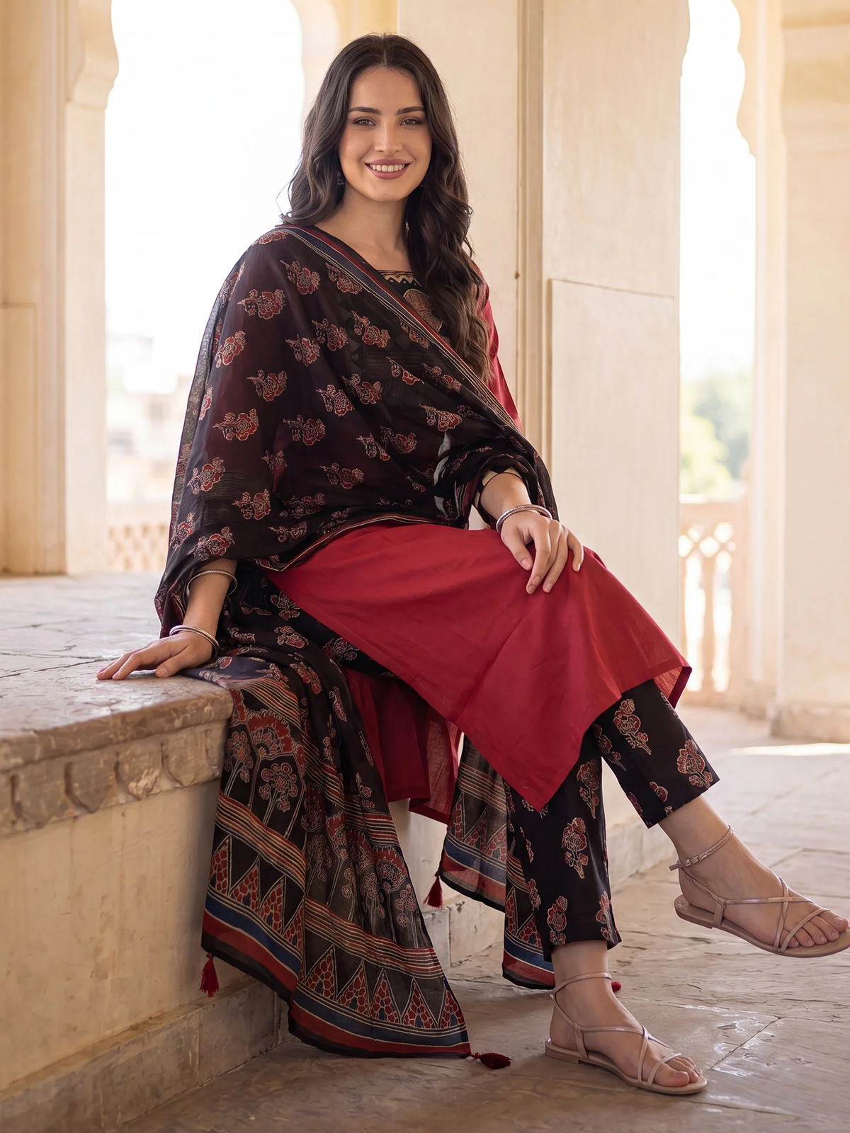 A woman models a traditional Indian salwar suit set consisting of a dark brown printed kurta and trousers, a solid red bottom fabric, and a matching red dupatta, seated in a stone corridor with arches.