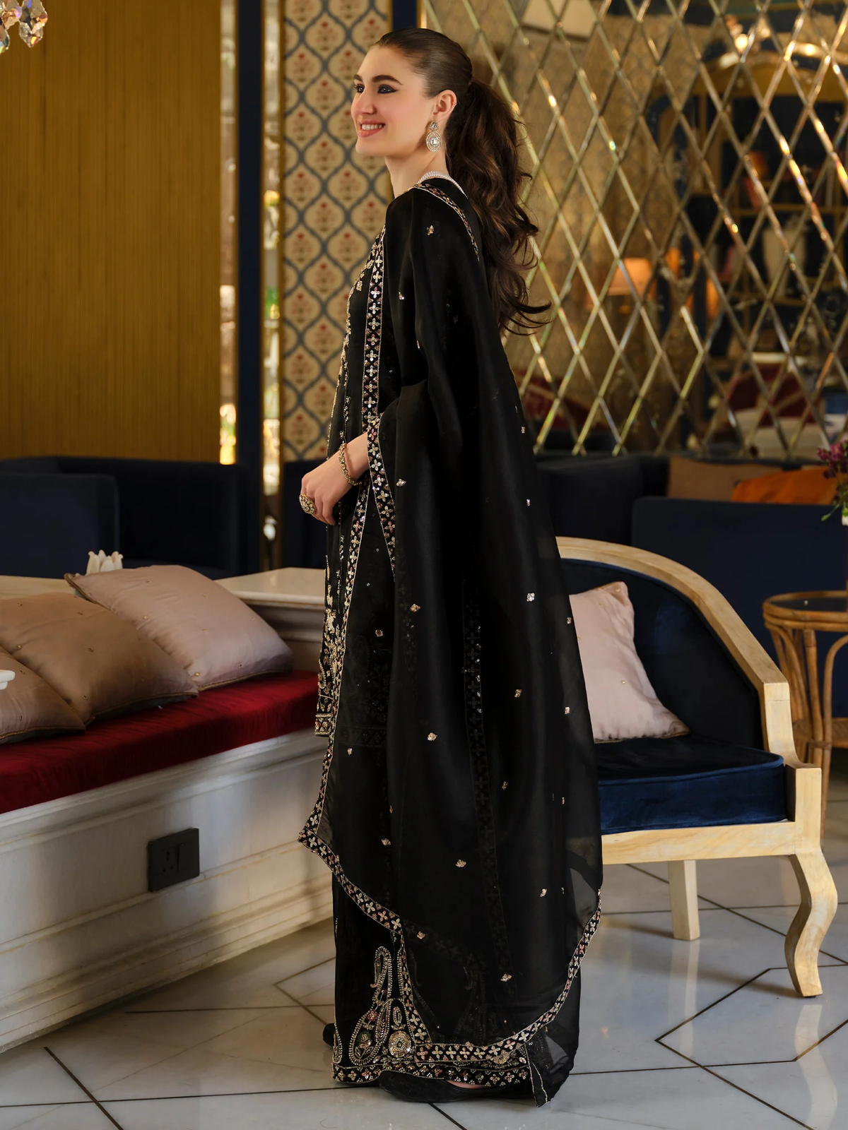 A woman wearing a black traditional outfit with intricate gold embroidery and a matching dupatta, standing in an indoor lounge setting with blue sofas and a patterned wall.
