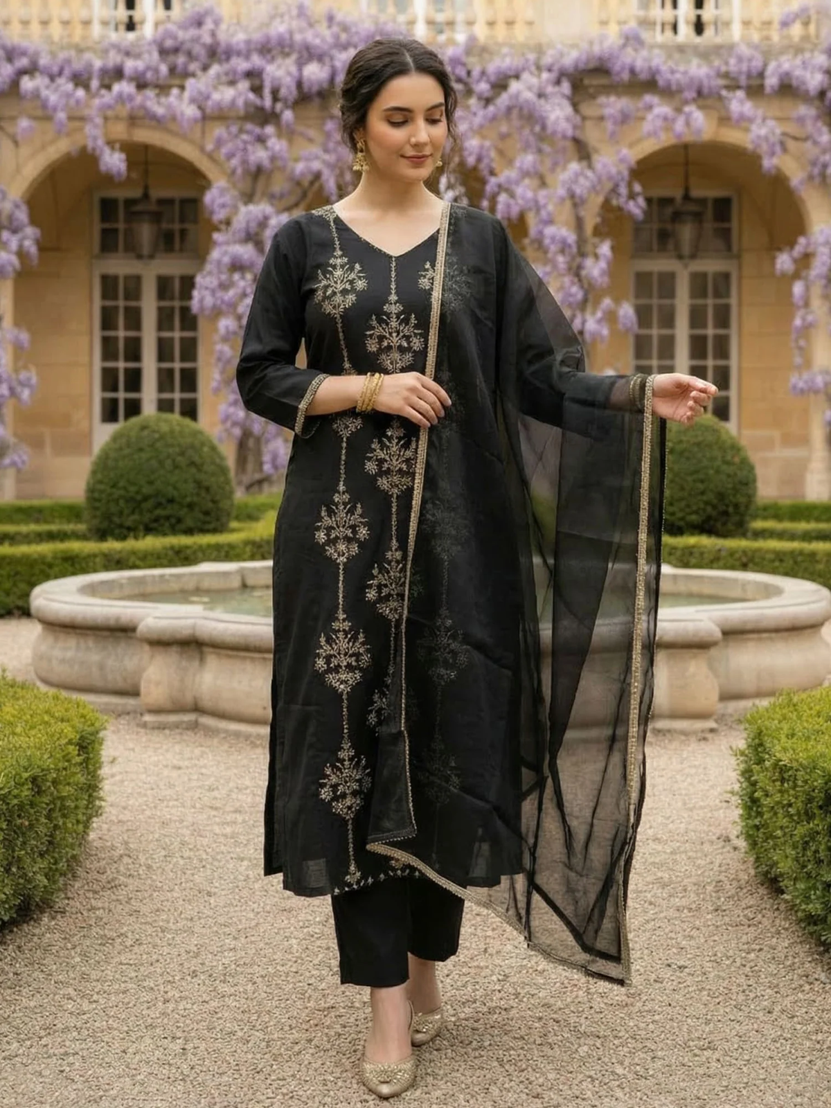A woman models a black three-piece ethnic suit with silver floral embroidery, consisting of a kurta, trousers, and a sheer dupatta, set against a garden backdrop with purple wisteria flowers.
