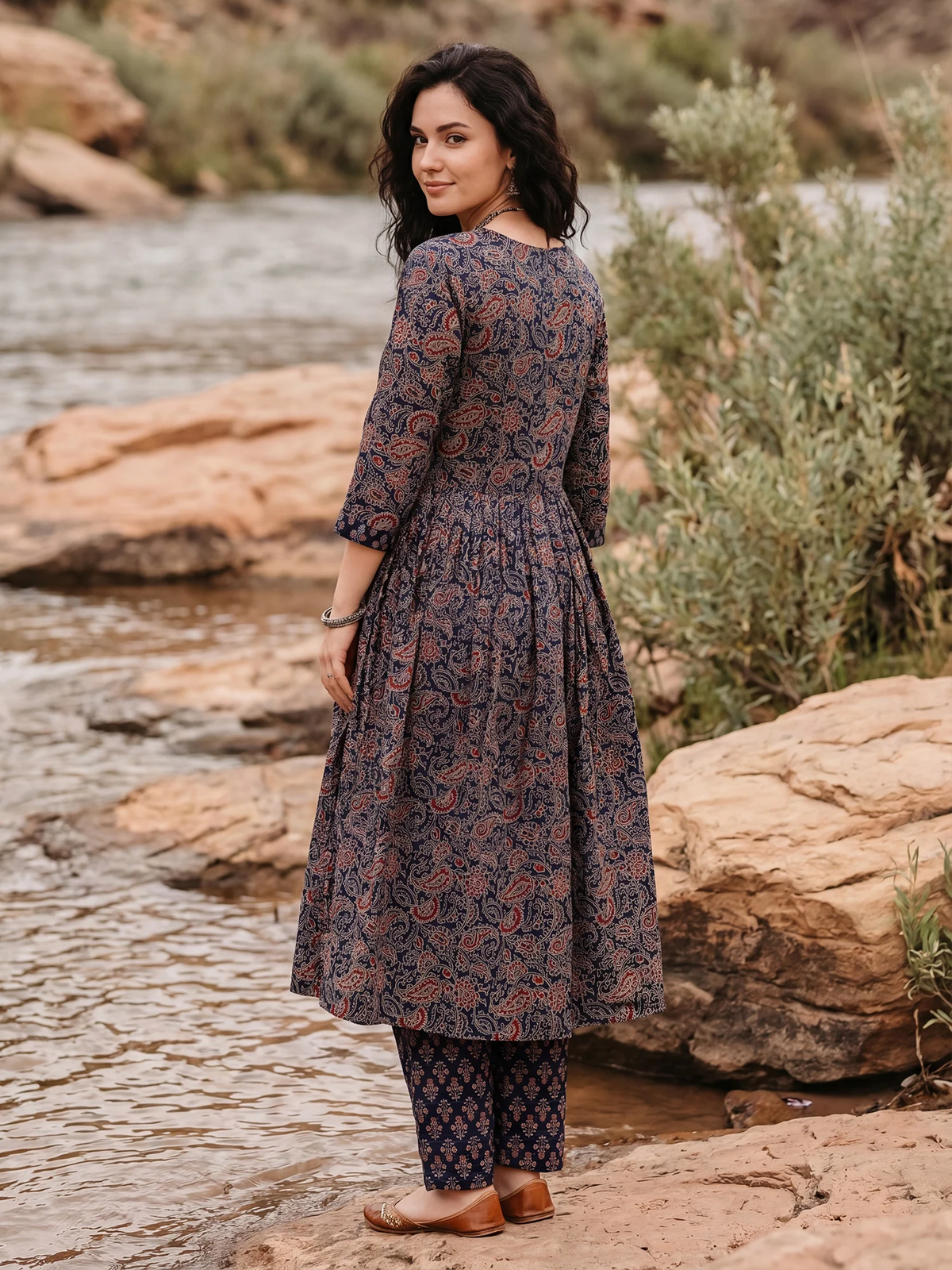 A woman stands outdoors by a rocky stream wearing a long-sleeve, knee-length printed tunic with matching patterned pants. The outfit has a dark base with a warm rust-orange and muted blue floral/paisley print.
