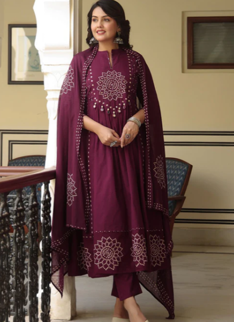 A woman models a traditional Indian outfit consisting of a deep purple kurta with white floral embroidery, matching trousers, and a matching sheer dupatta draped over her shoulders, set against an indoor architectural background.