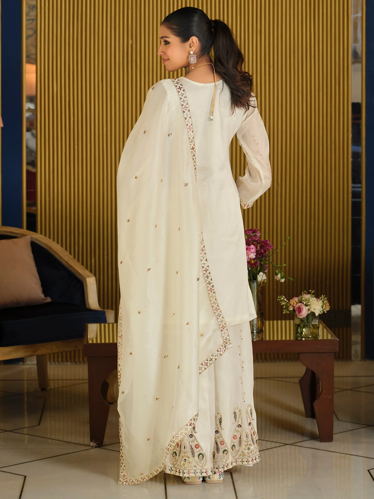 Woman in a white embroidered outfit standing in an indoor setting with decor elements.