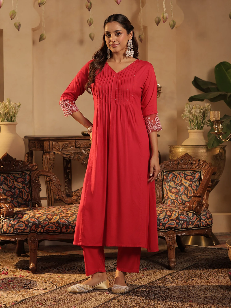 Woman in a red traditional outfit standing in an ornate room with decorative furniture and plants.