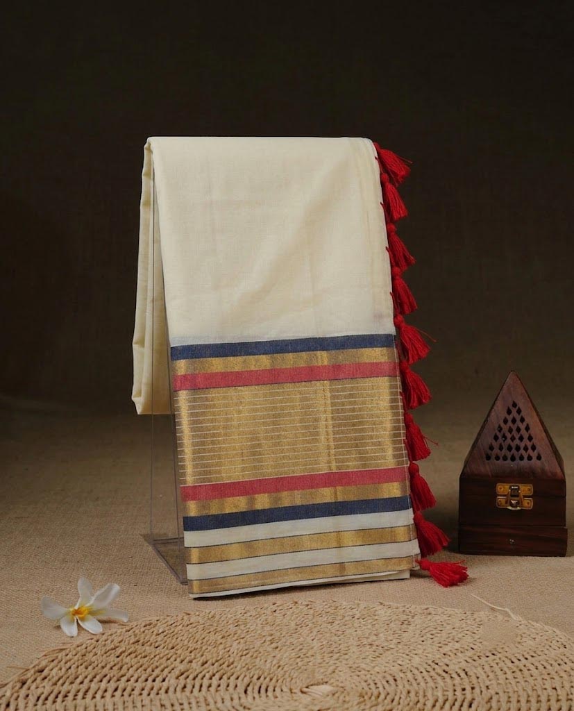 A folded cream-colored silk saree draped on a clear stand, featuring a gold zari border with red, blue, and white stripes, and red tassels along the edge. The saree is styled on a beige surface with a small white flower and a dark wooden box.