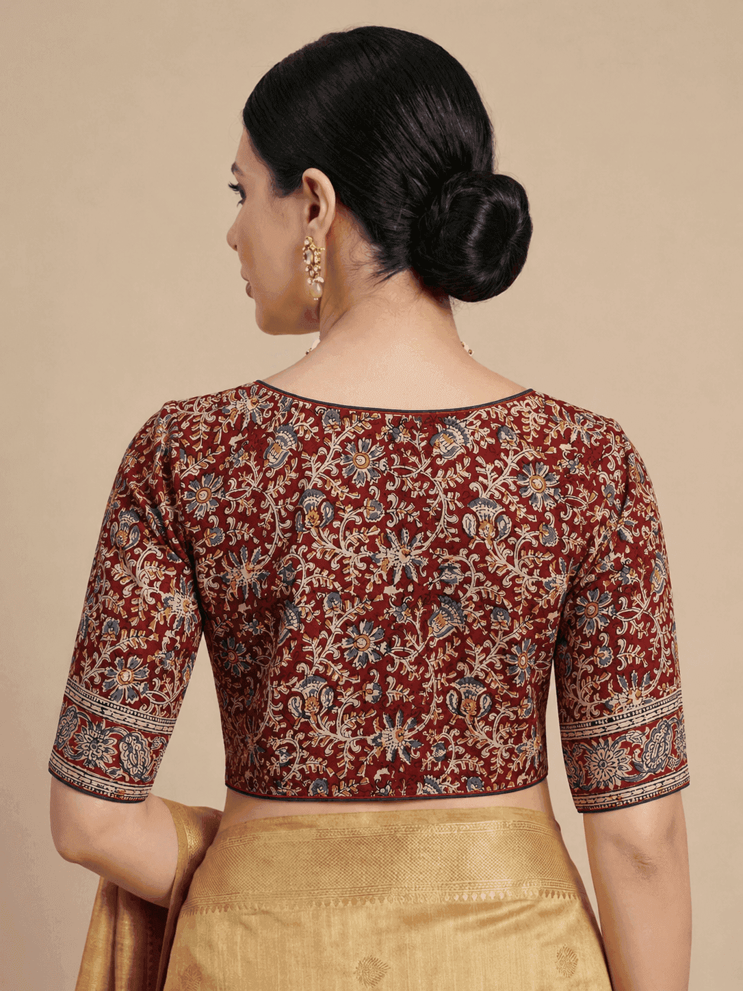 A woman shown from the back wearing a maroon printed blouse with beige, blue, and gold floral motifs, paired with a gold skirt, against a solid beige background.