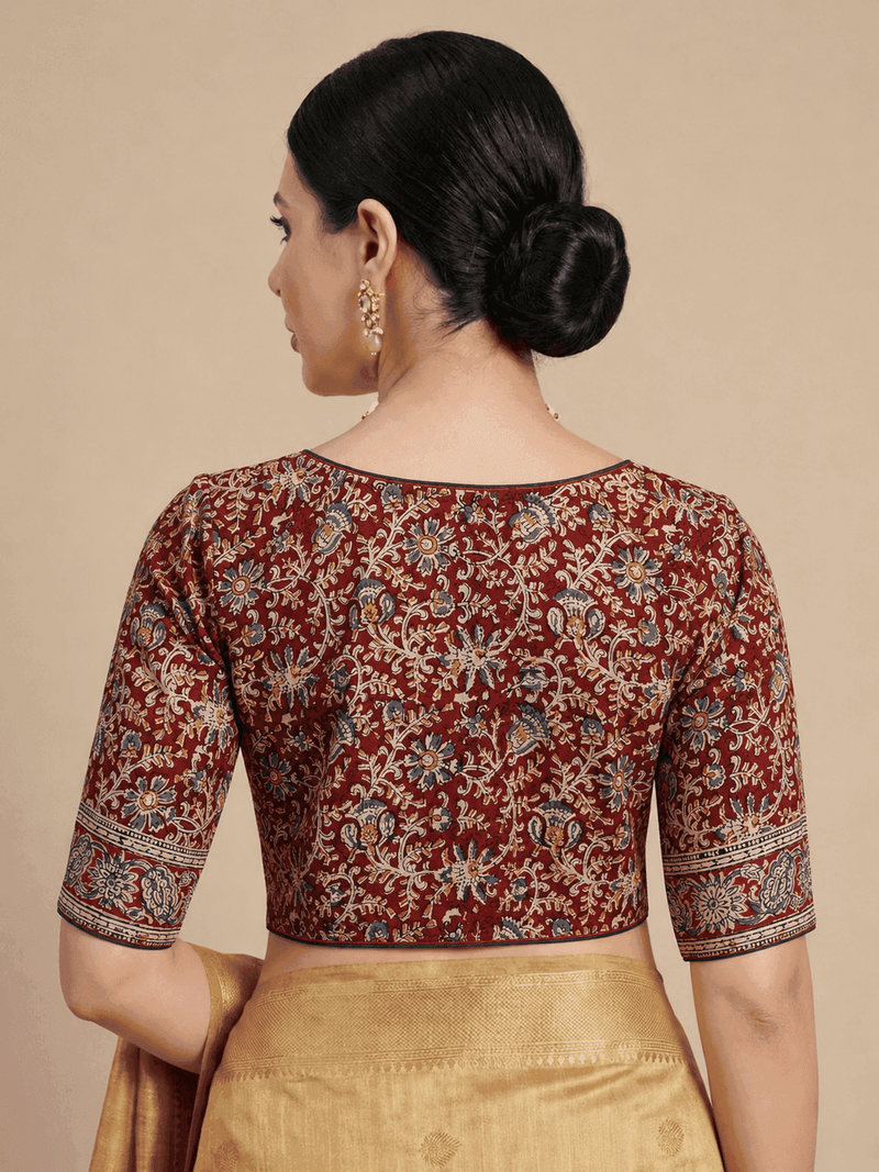 A woman shown from the back wearing a maroon printed blouse with beige, blue, and gold floral motifs, paired with a gold skirt, against a solid beige background.