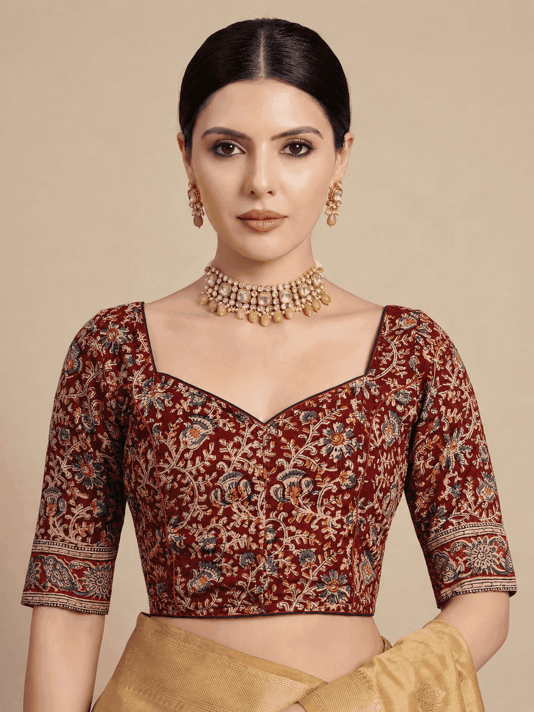A woman modeling a deep maroon red printed cotton blouse with intricate gold, beige, and blue floral patterns, paired with a gold necklace and earrings.