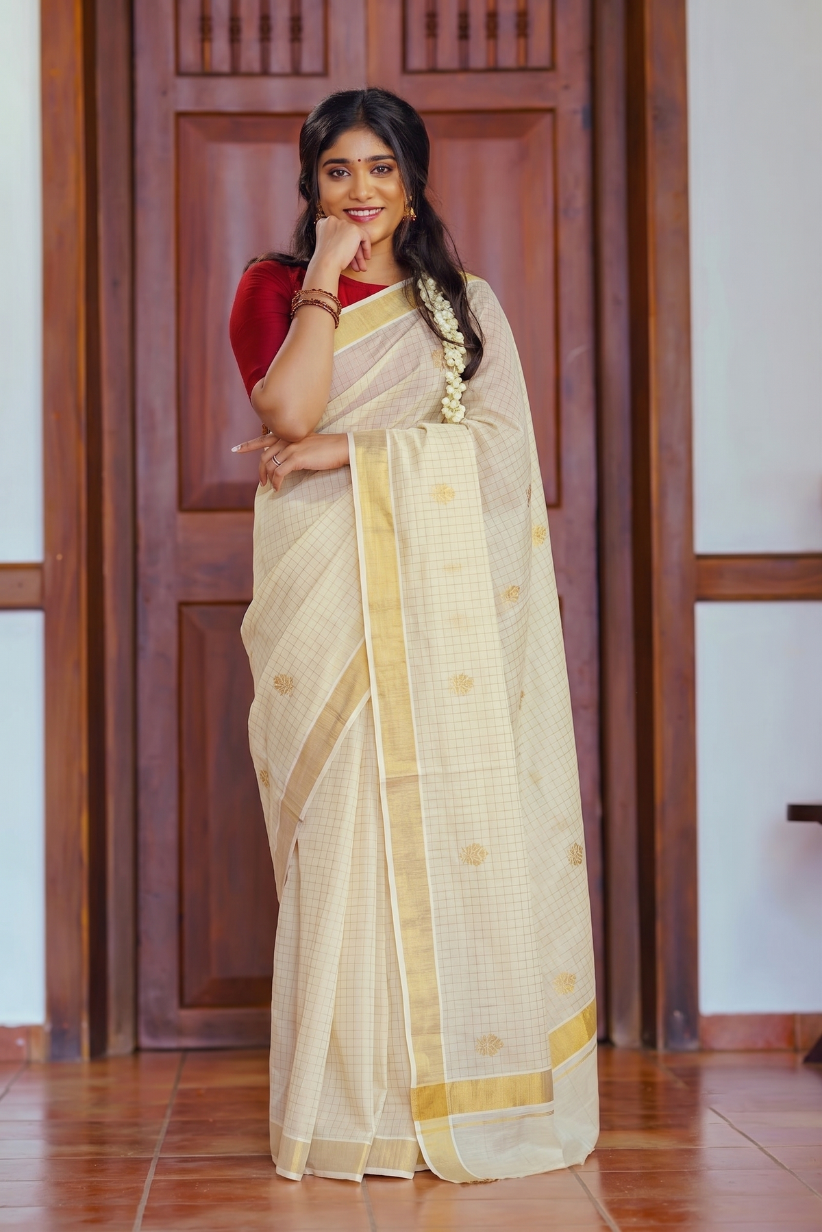 A woman wearing a traditional Indian saree stands in front of a wooden door. The saree is cream-colored with a subtle grid pattern, gold border stripes, and small gold motifs, paired with a red blouse.