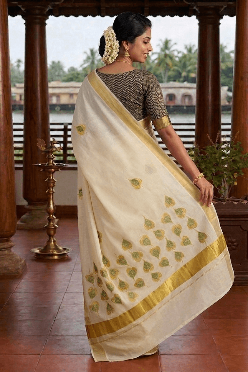 A woman wearing a traditional Indian saree, photographed from behind as she walks through a wooden corridor. The saree is off-white with gold borders and green leaf motifs, paired with a patterned gold and black blouse.