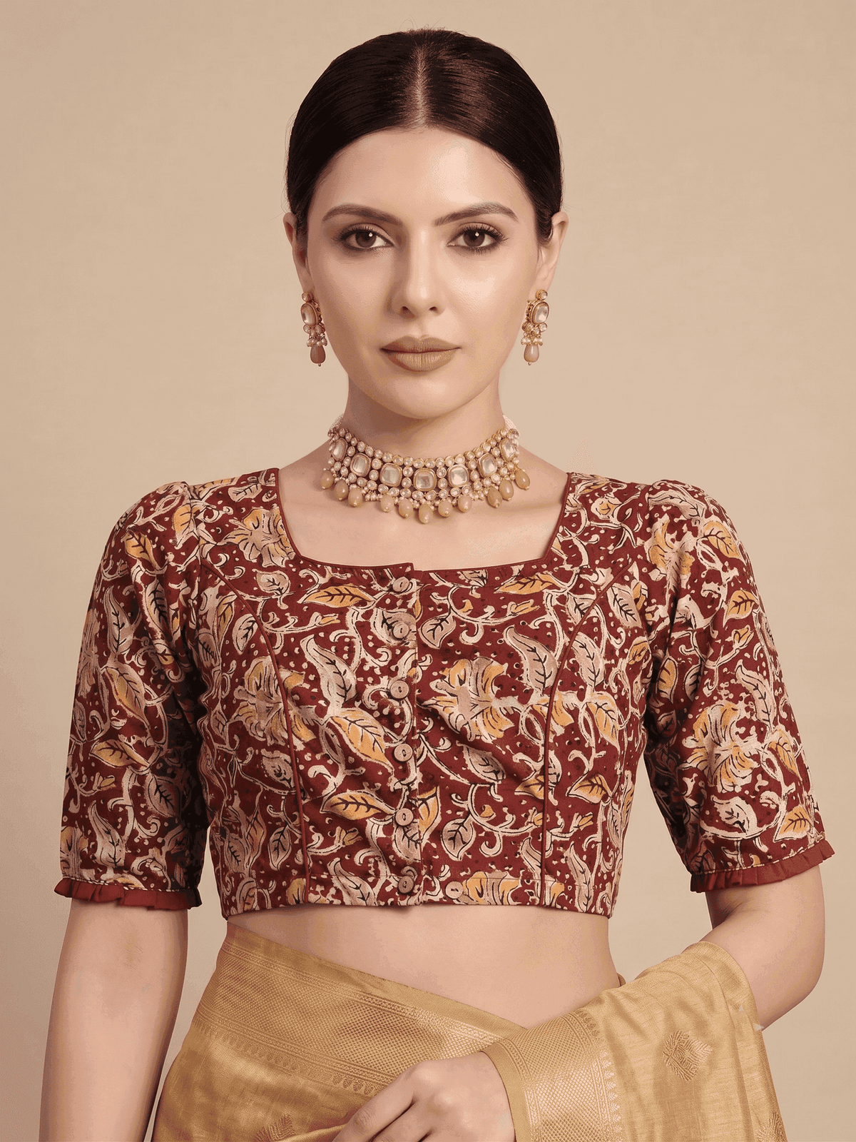 A woman modeling a short-sleeved crop blouse with a maroon and gold paisley pattern, paired with a gold skirt and jewelry, against a solid beige background.