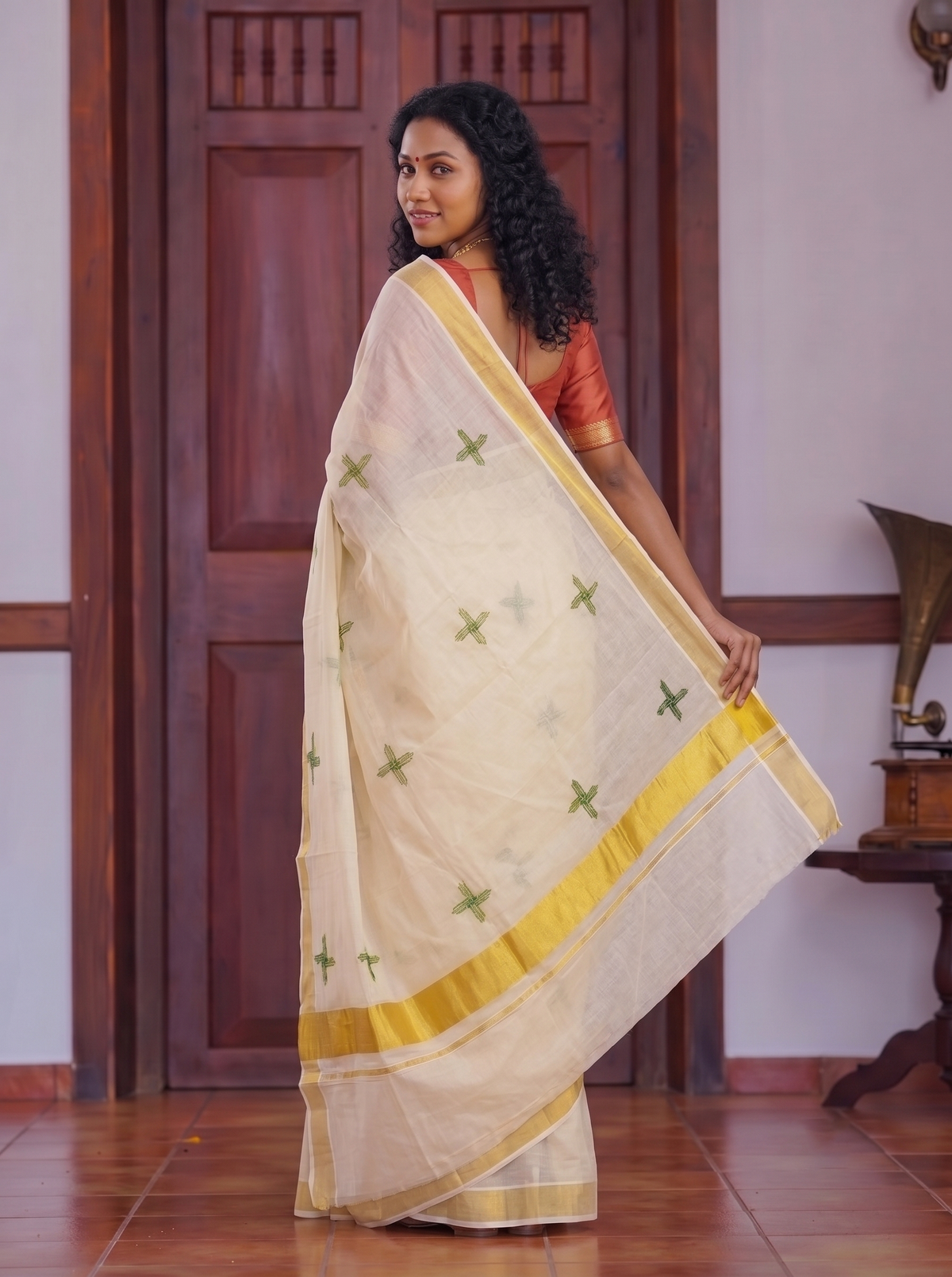 A woman wearing a cream-colored saree with green cross motifs and a yellow border, standing in front of a dark wooden door in an indoor setting.