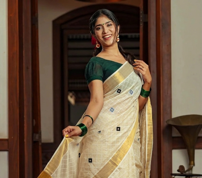 A woman wearing a cream-colored saree with gold borders and a dark green blouse, standing in a doorway with wooden paneling.