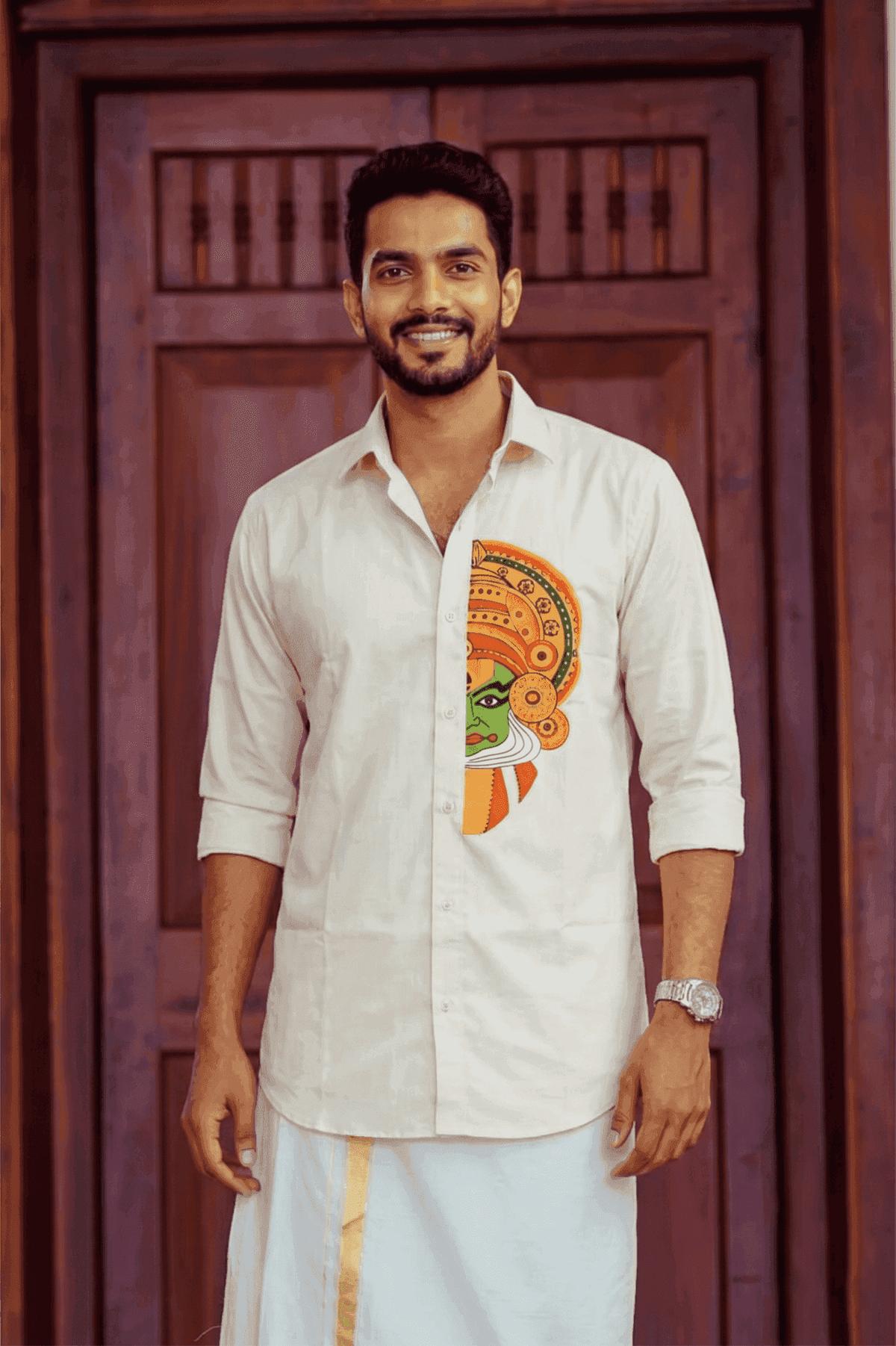 A man wearing a light beige Tamil Nadu style kurta with a colorful traditional face print on the front, paired with a white dhoti, standing in front of a dark wooden door.