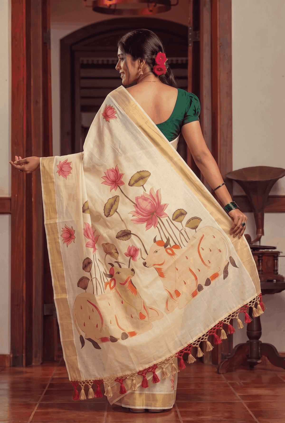 A woman wearing a cream-colored saree with pink lotus flowers, green leaves, and a gold border, paired with a green blouse, standing in a traditional indoor setting.