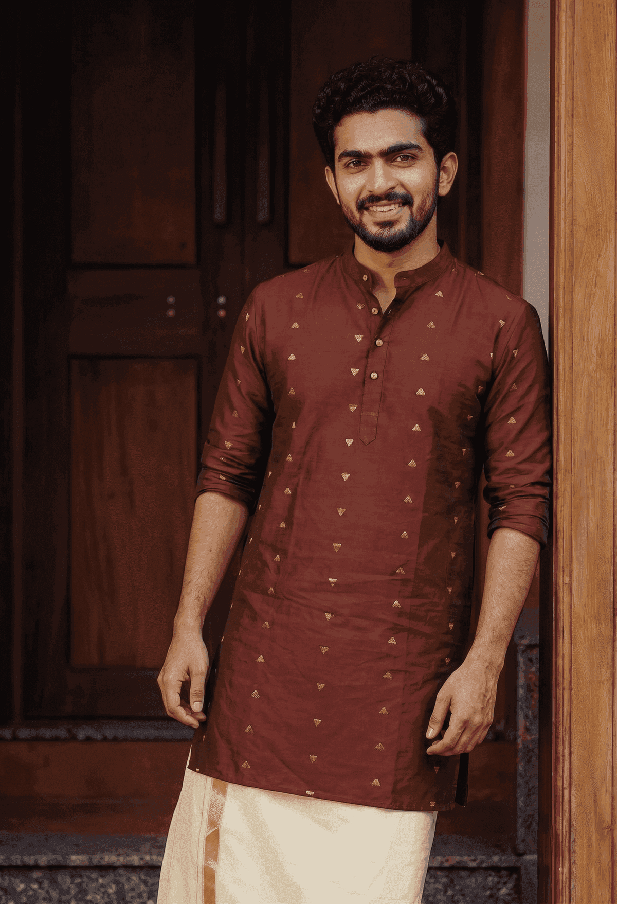 A man wearing a rust-brown long-sleeve kurta with small gold motifs and a cream dhoti, standing in front of a dark wooden door.