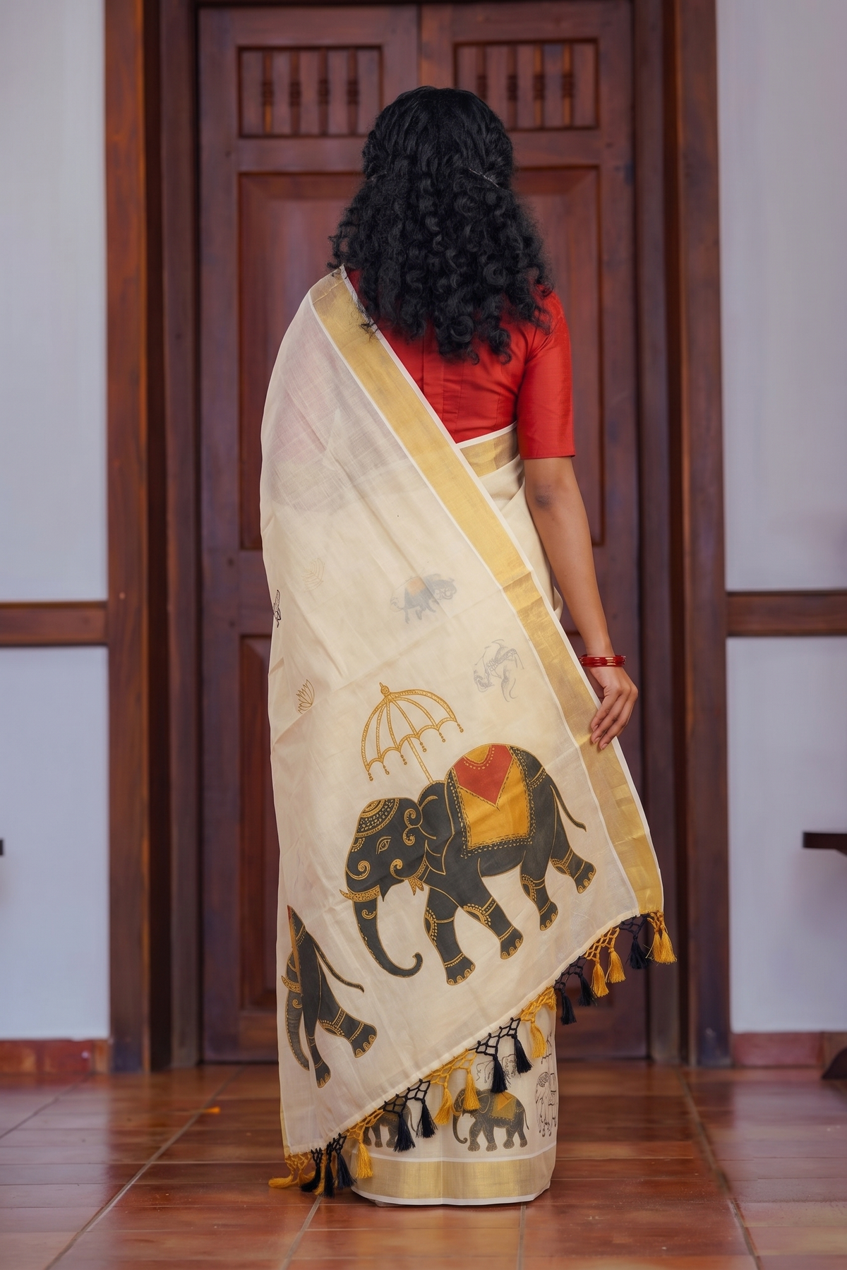 A woman viewed from behind wearing a cream-colored cotton saree with elephant prints and a red blouse, standing in front of a dark wooden door.