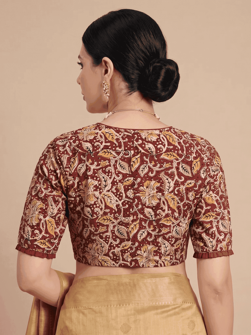 A woman shown from the back wearing a short-sleeve printed blouse in rust red, beige, and orange with a gold skirt, set against a solid beige background.