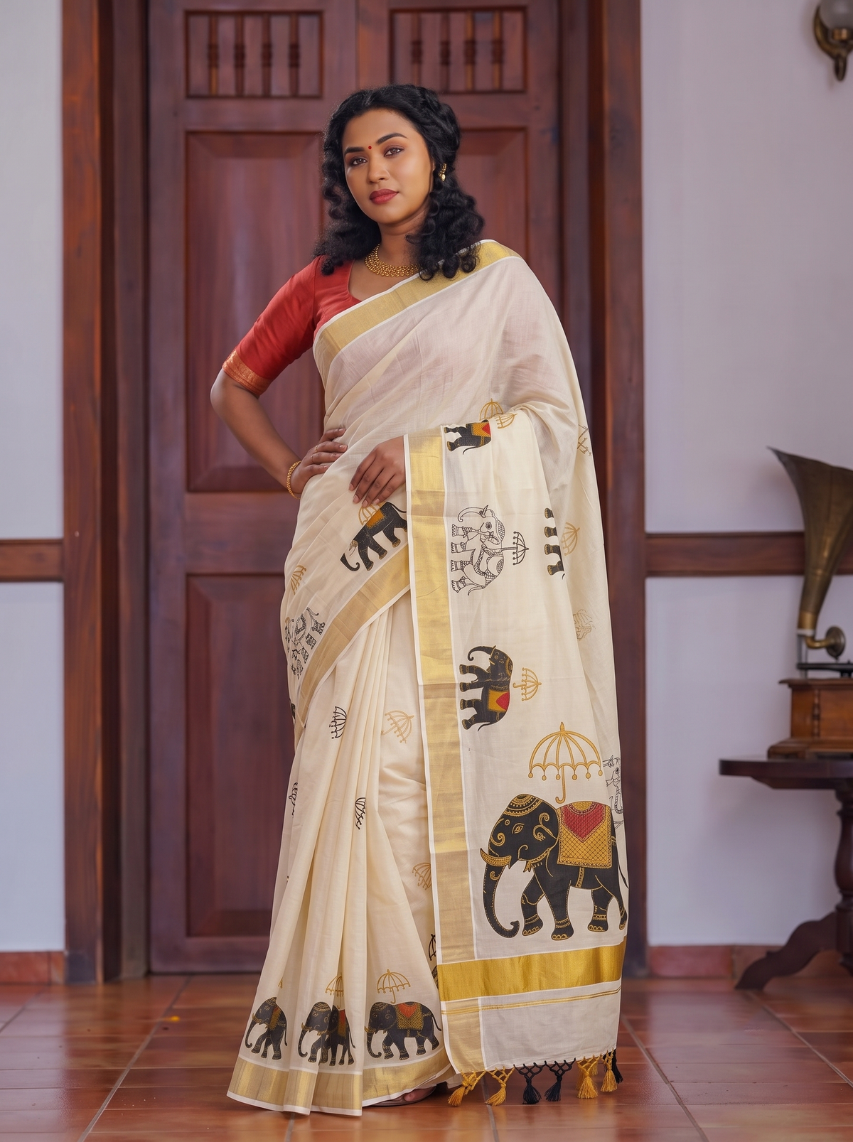 A woman models a cream-colored cotton saree with a red sleeveless blouse. The saree features printed motifs of elephants, a palmyrah tree, and a gold border, with a matching cream dupatta draped over her shoulder.