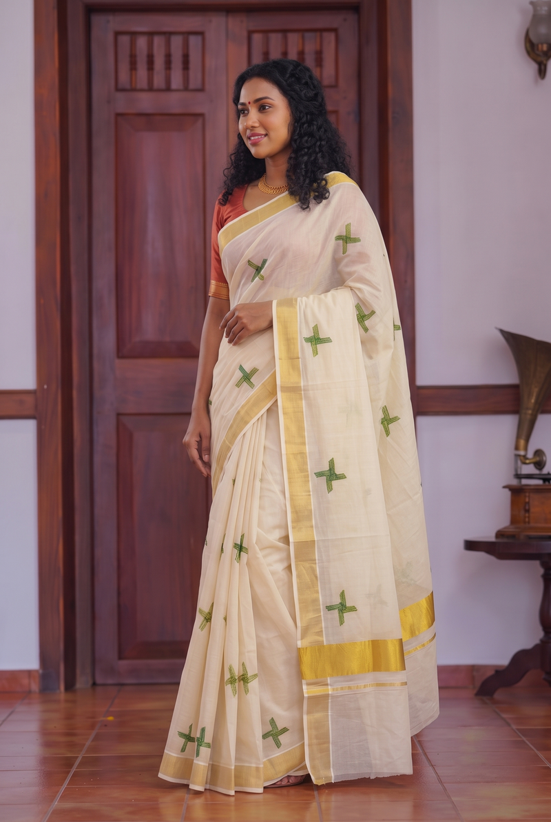 A woman wearing a cream-colored silk saree with green cross motifs and yellow borders, paired with a red sleeveless blouse, standing in front of a wooden door.
