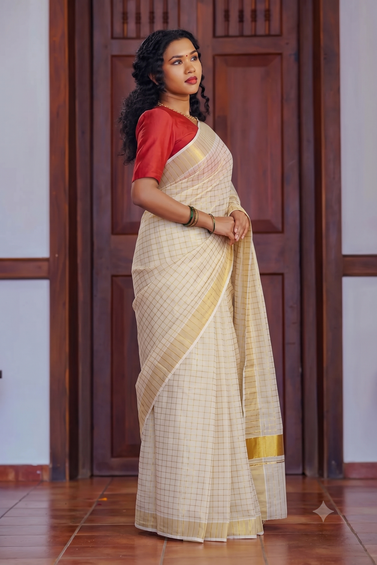A woman modeling a traditional Indian saree in a warm cream and beige checkered pattern with a red short-sleeve blouse, standing in front of a dark wooden door.