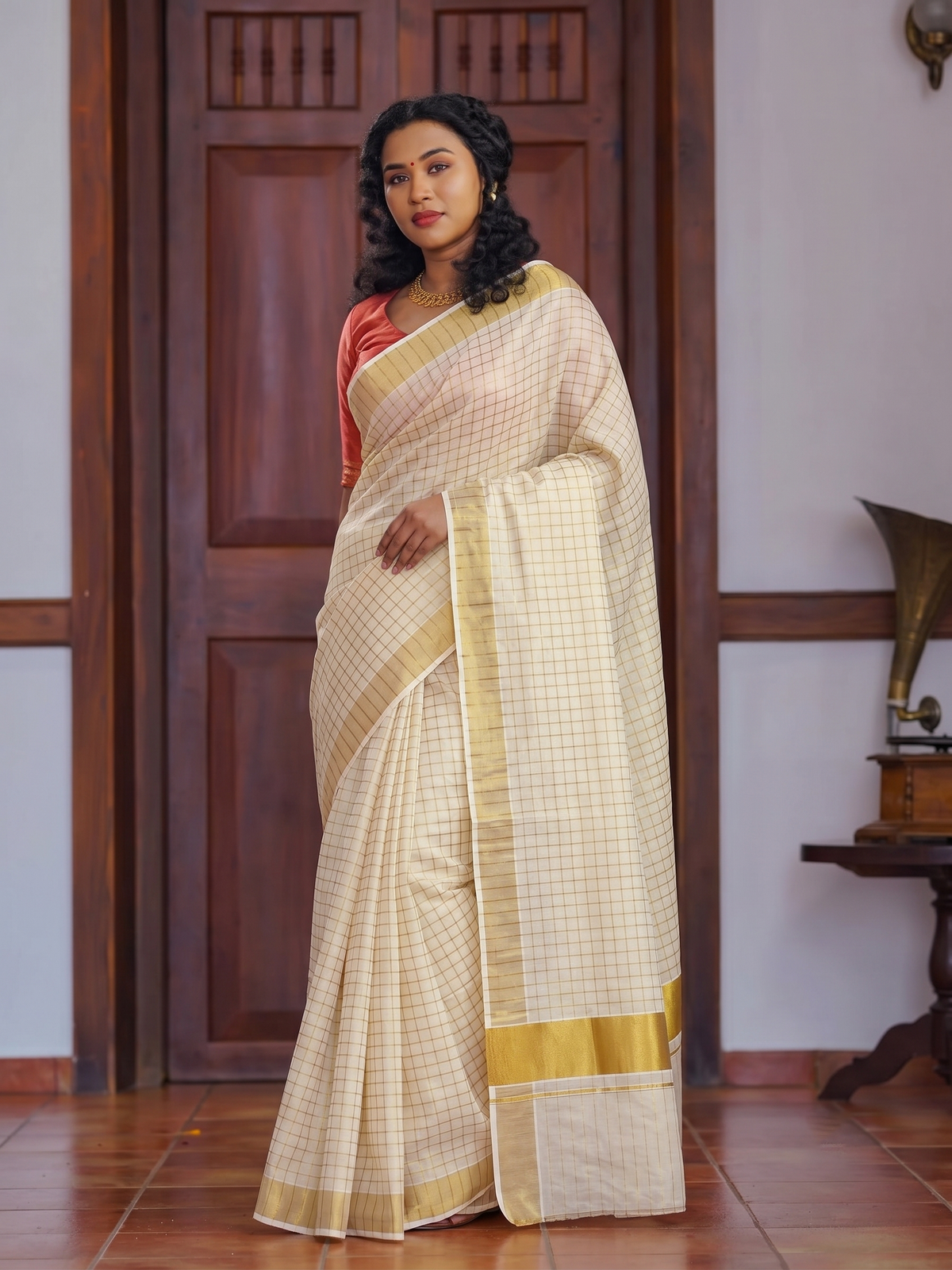 A woman models a traditional Indian saree in a warm cream-beige color with a subtle grid pattern and a wide gold border, paired with a coral-pink blouse, set against a background of a dark wooden door and tiled floor.