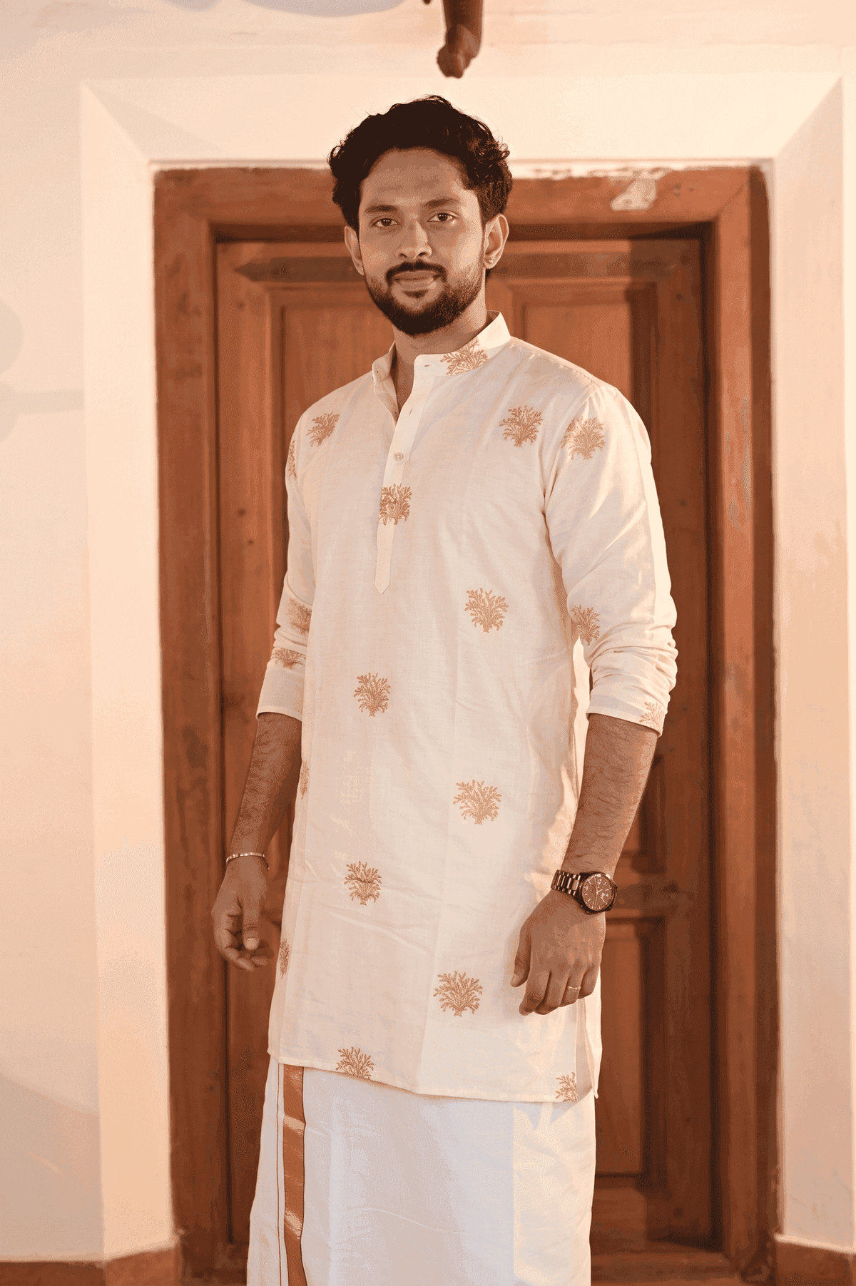 A man wearing a traditional Indian outfit consisting of a cream-colored kurta with gold floral embroidery and a white dhoti, standing in front of a wooden door.