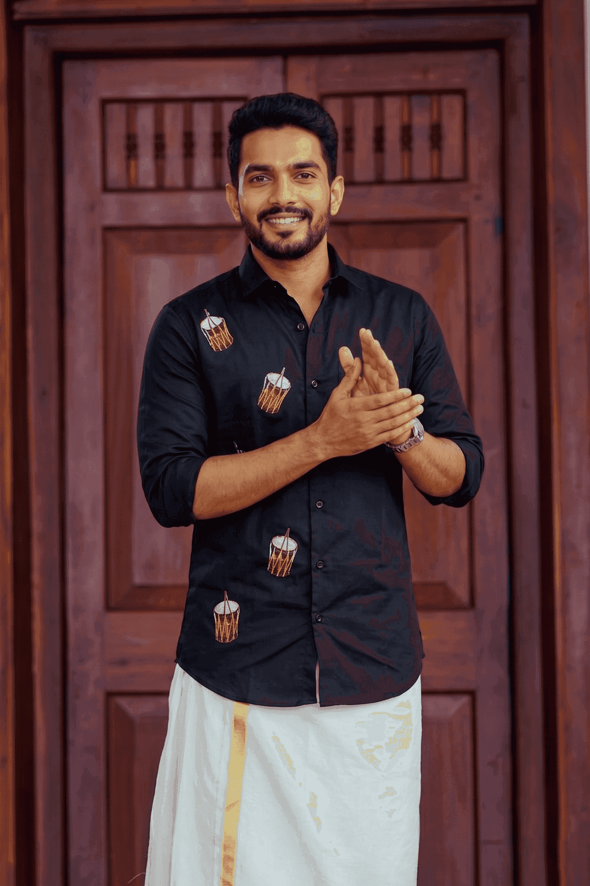 A man wearing a black long-sleeve button-up shirt with embroidered drum motifs and a white traditional dhoti with a gold stripe, standing in front of a dark wooden door.