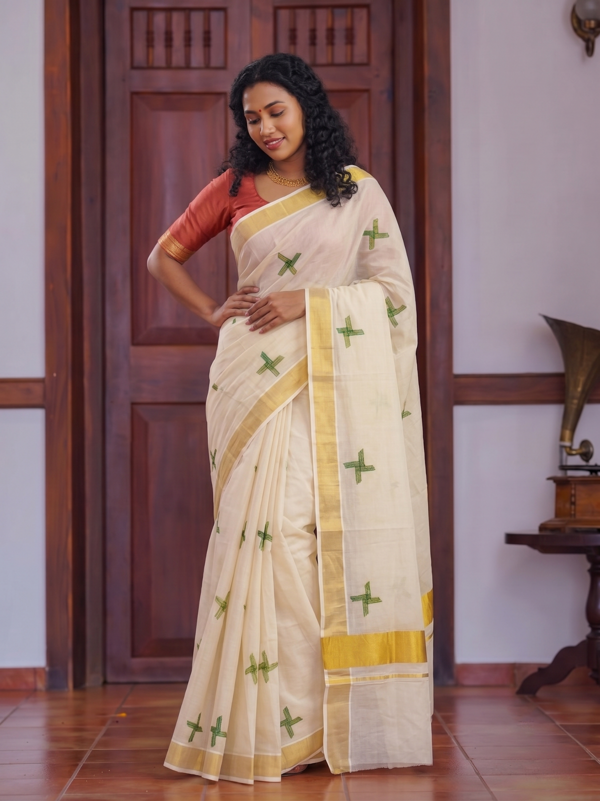A woman modeling a cream-colored cotton saree with green cross motifs and a yellow border, paired with a rust-orange blouse, standing in front of a dark wooden door.