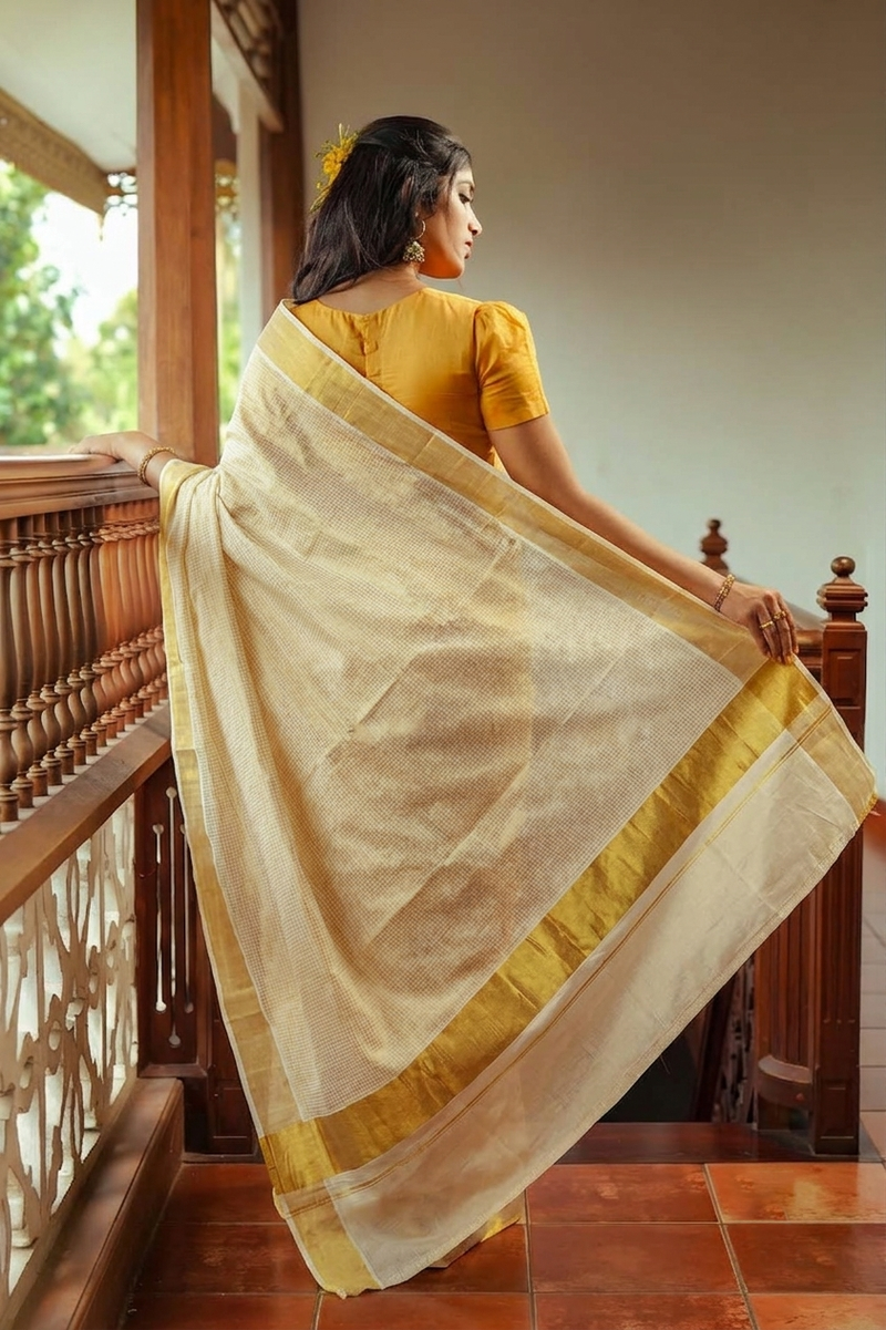 A woman stands on a wooden balcony wearing a cream-colored silk saree with a wide golden-yellow border and a mustard yellow blouse, holding the saree out to display its drape.